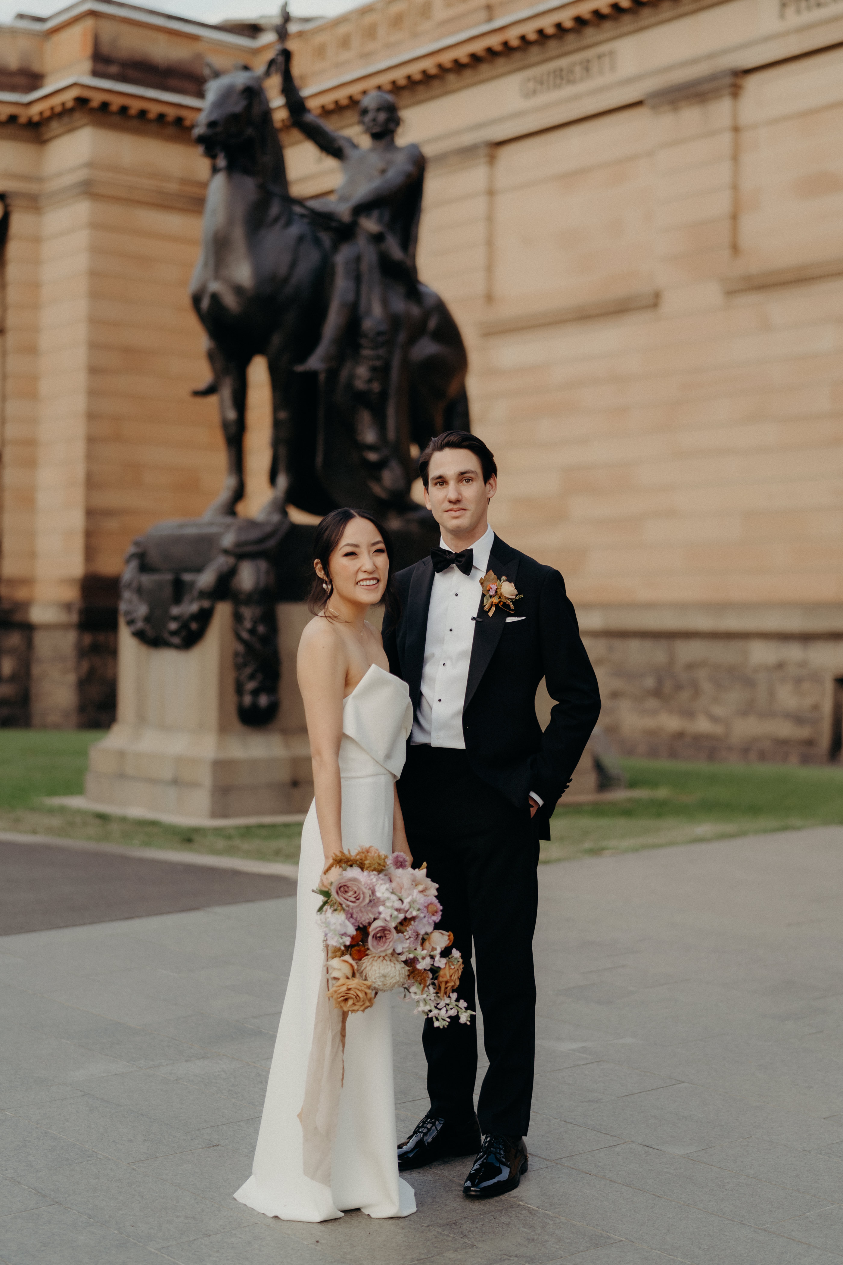 A person in a suit and a person in a wedding dress, carring a bouquet, stand in front of a large standstone building and a statue of a person on horseback.