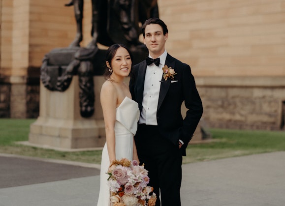 A person in a suit and a person in a wedding dress, carring a bouquet, stand in front of a large standstone building and a statue of a person on horseback.