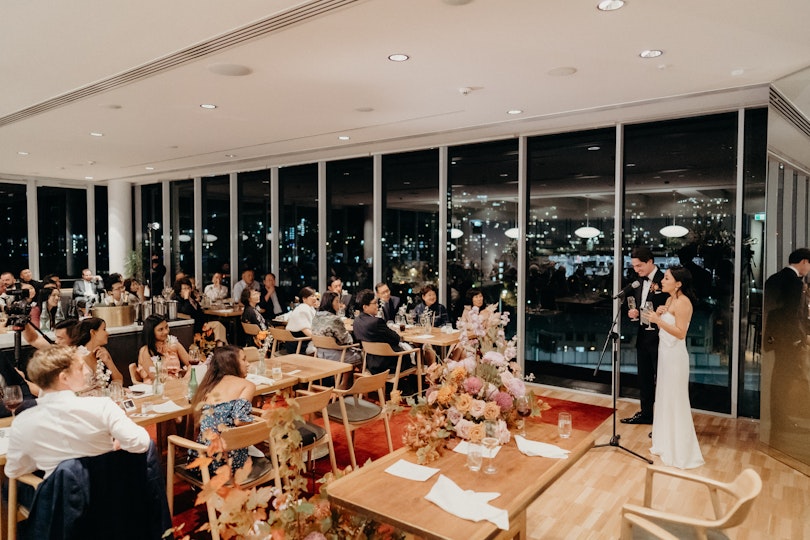 A couple stand at a microphone in front of tables of seated people. City lights at night are visible through floor-to-ceiling windows.