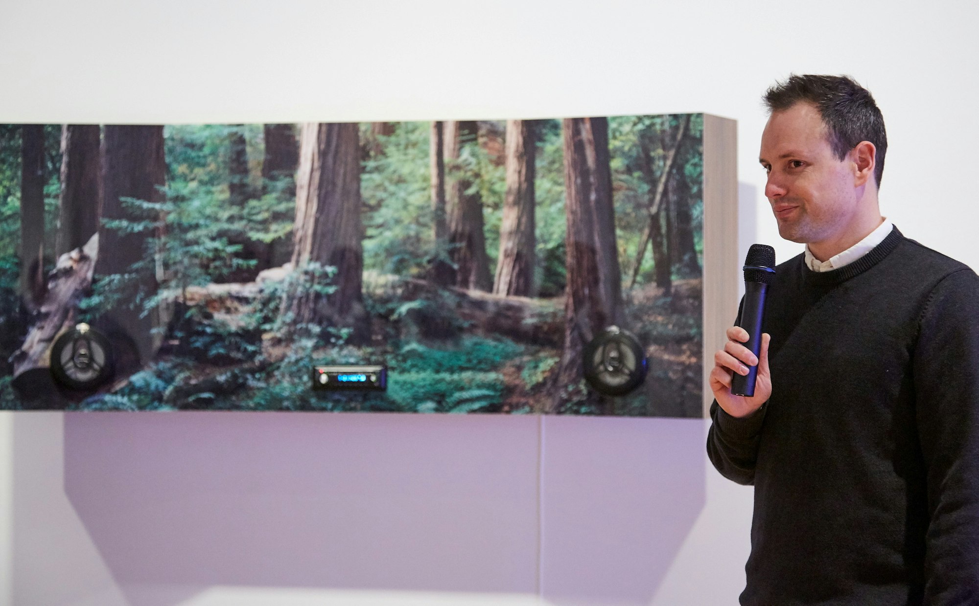 A man with short dark hair, dressed in a white shirt and black jumper, is holding a microphone. To his right is a long narrow panel with in-built audio speakers  and a photo of forest trees, taken at ground level.