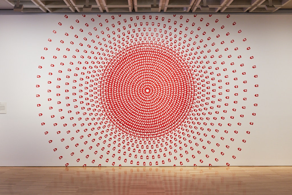 Bright red tags each with a white letter S, A, L or E, arranged in concentric circles on a wall. The tags in the inner circles overlap, while those in the outer circles are increasingly spaced.