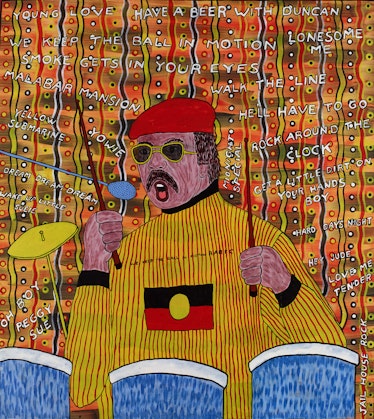 A man at a drumkit with his drumsticks raised. He has brown hair and a moustache, and is wearing a beret, sunglasses and a long-sleeved top with the Aboriginal flag on the chest. The titles of songs are written on the patterned background.