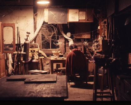 A cluttered room, lit by a single ceiling light, in which a woman sits bent over a work table full of small objects