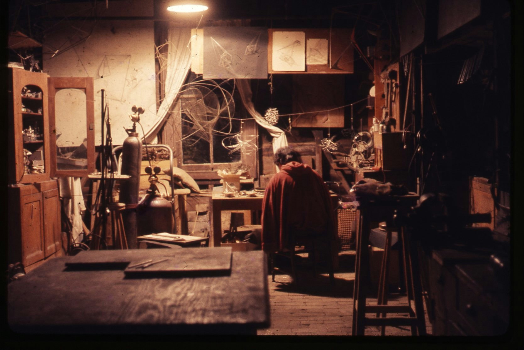 A cluttered room, lit by a single ceiling light, in which a woman sits bent over a work table full of small objects