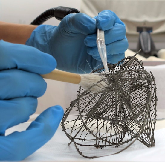 Blue-gloved hands work with a small vacuum nozzle and a brush on a small wire sculpture.