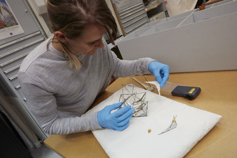 A conservator wearing blue gloves holds a small boomerang-shaped object in her left hand. Her right hand rests on a cushion on which there is a similar object and a small sculpture.