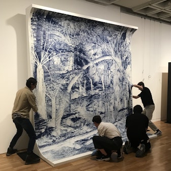 Four people move a large blue-and-white drawing on a curved white support into position against a gallery wall.