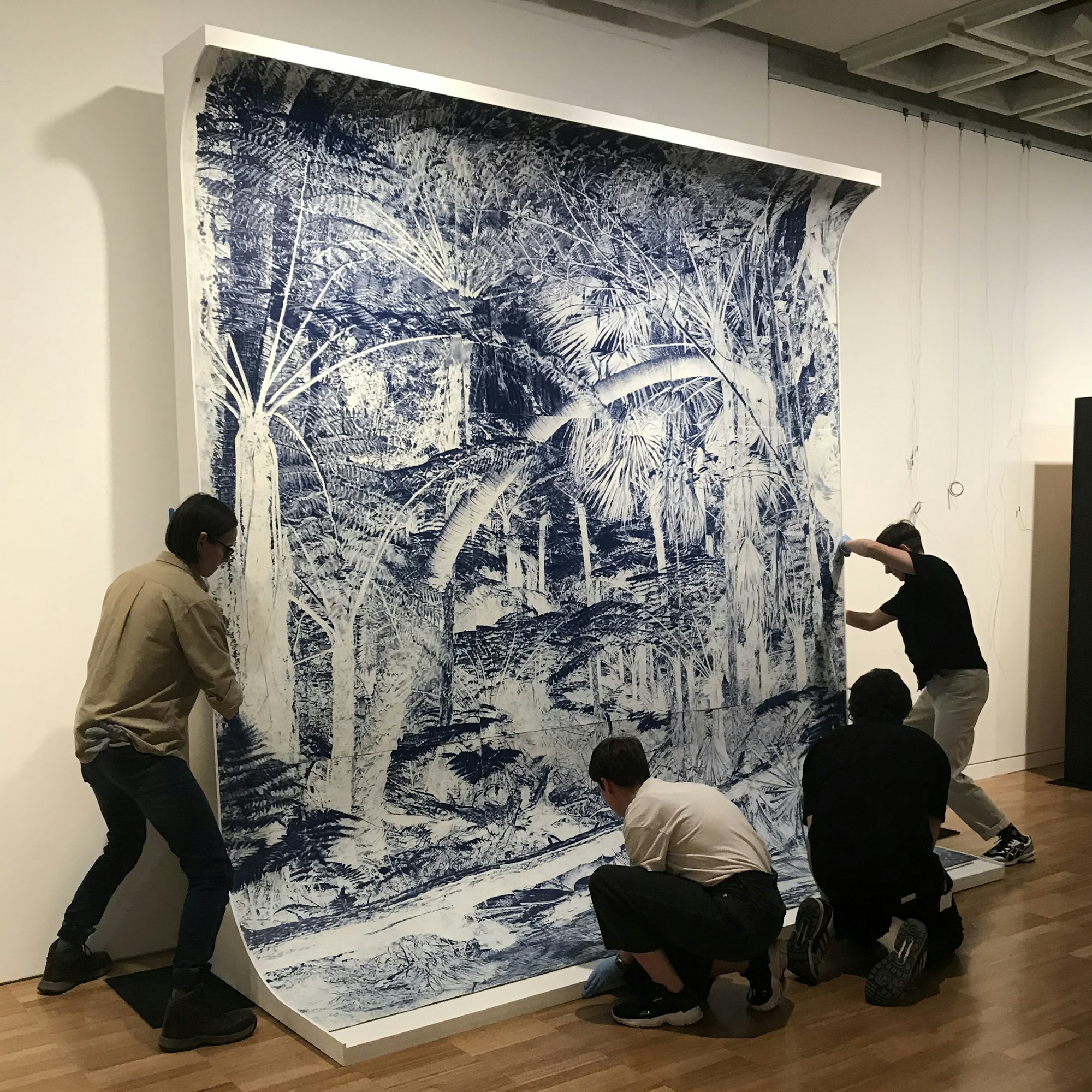 Four people move a large blue-and-white drawing on a curved white support into position against a gallery wall.