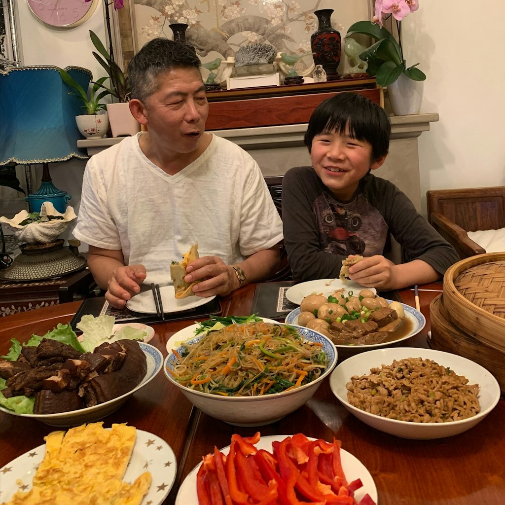 A Chinese man and a boy sit at a dining table laden with dishes of food. Each as a half-eaten wrap in his left hand.