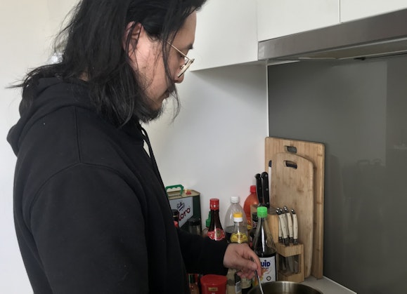 A dark-haired and bearded man wearing glasses and a black hoodie stirs something in a saucepan on a stove. Next to him are chopping boards, knives and various bottles.