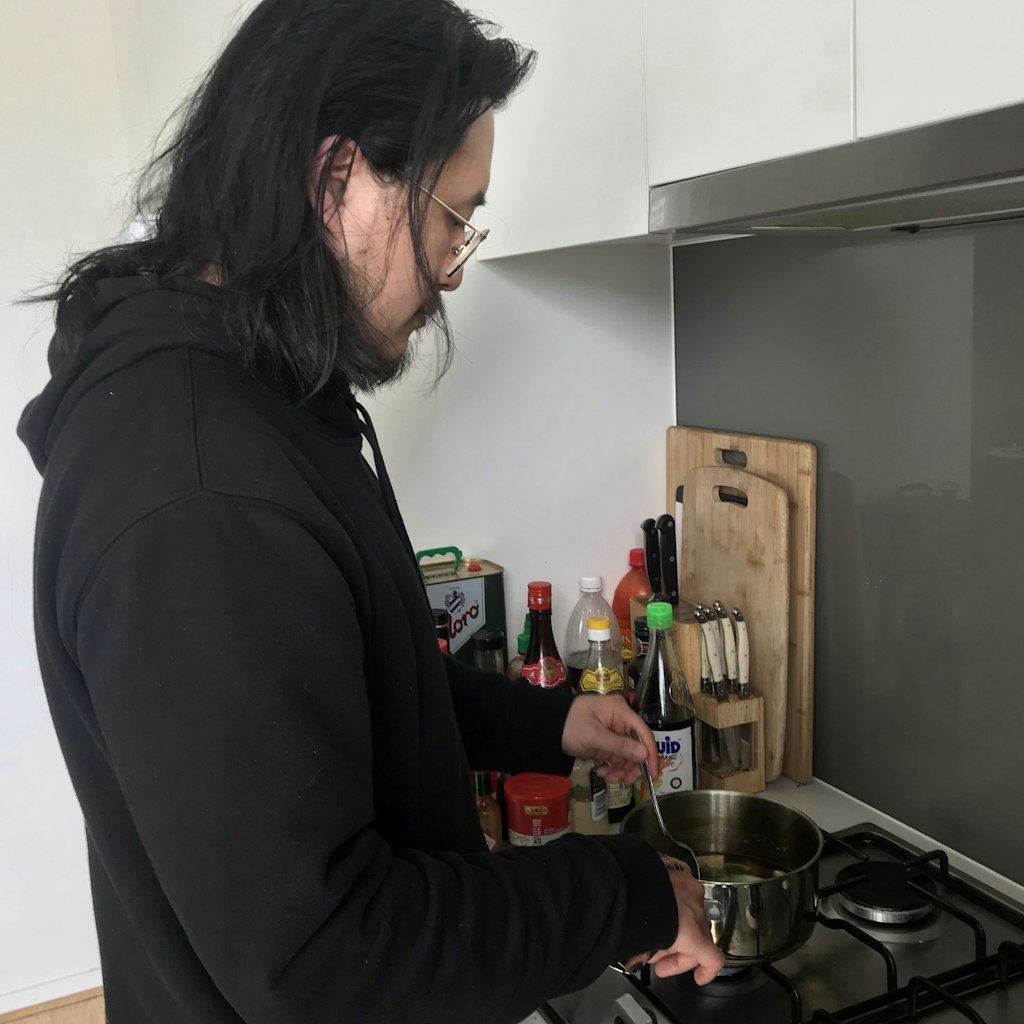 A dark-haired and bearded man wearing glasses and a black hoodie stirs something in a saucepan on a stove. Next to him are chopping boards, knives and various bottles.