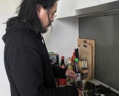 A dark-haired and bearded man wearing glasses and a black hoodie stirs something in a saucepan on a stove. Next to him are chopping boards, knives and various bottles.