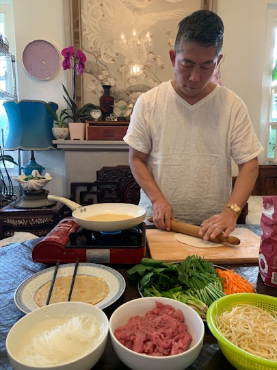 A man with short dark hair and a white t-shirt uses a rolling pin to make a pancake. On the table on which he works are other ingredients and a small gas stove.