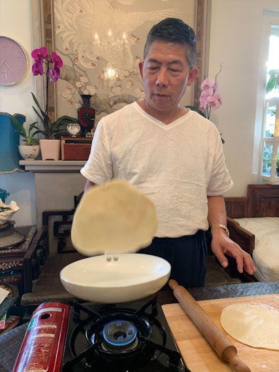 A man with short dark hair and a white t-shirt flips a pancake in a pan on small gas stove.