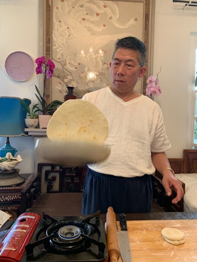A man with short dark hair and a white t-shirt flips a pancake in a pan on small gas stove.