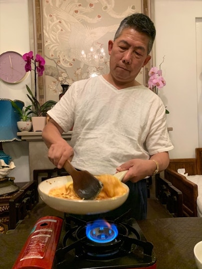 A man with short dark hair and a white t-shirt stir-frying in a pan on small gas stove.