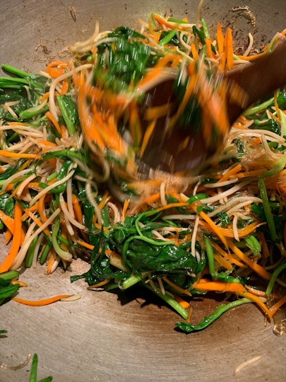 A mix of spinach leaves, carrot strips and soybean sprouts on the base of a wok, being stirred with a wooden spoon.