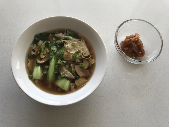 White bowl containing soup with pieces of bok choy, tofu and mushrooms and a small glass blow containing pickles.