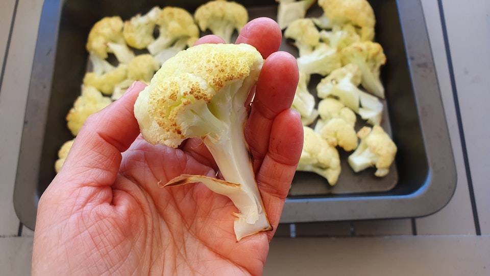 A hand holding a cauliflower floret with a baking dish holding other cauliflower pieces in the background.