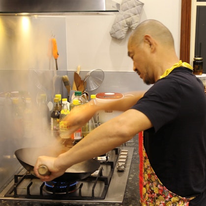 A bearded, shaven-headed man in a black t-shirt and floral apron cooking in a wok on a gas stove. There are various bottes and cooking implements in the background.