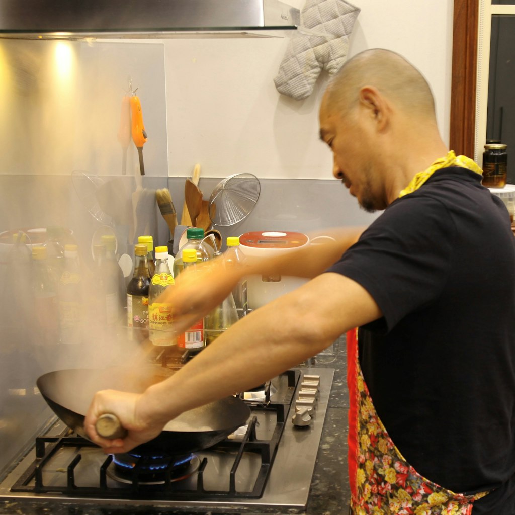 A bearded, shaven-headed man in a black t-shirt and floral apron cooking in a wok on a gas stove. There are various bottes and cooking implements in the background.