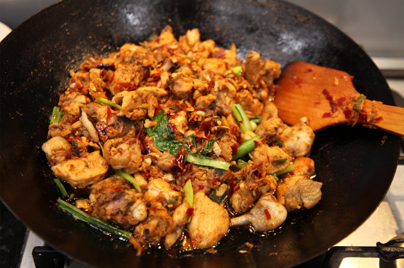 A mix of chicken pieces, chili, greens and other ingredients being stirred in a wok with a wooden spoon.