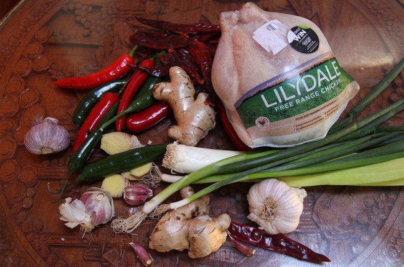 Garlic, ginger, chillies, spring onion, a leek and a whole chicken lie on a table.