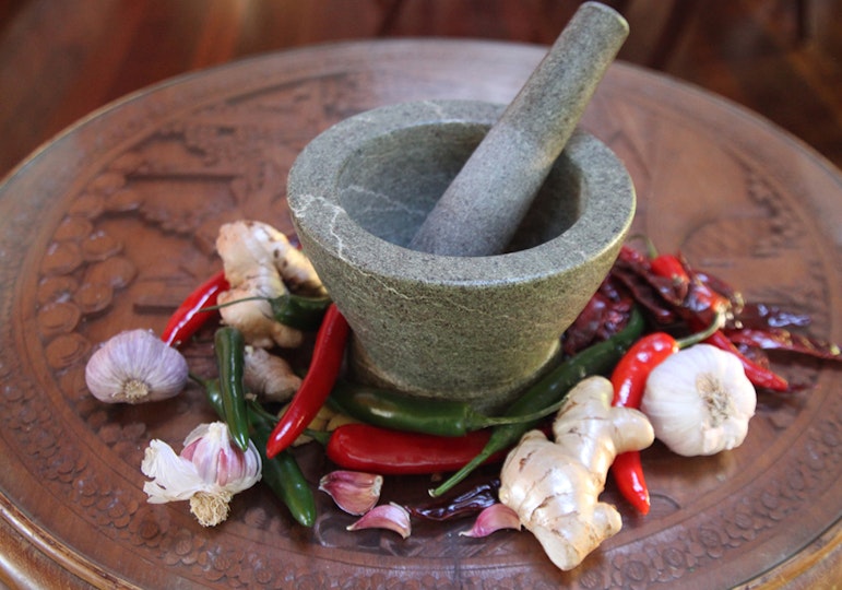 A mortar and pestle on a wooden table surrounded by ginger, garlic and chillies.