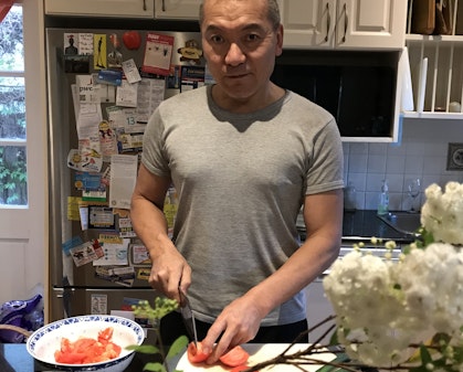 A man with short dark hair and a grey t-shirt cuts tomatoes at a kitchen bench.
