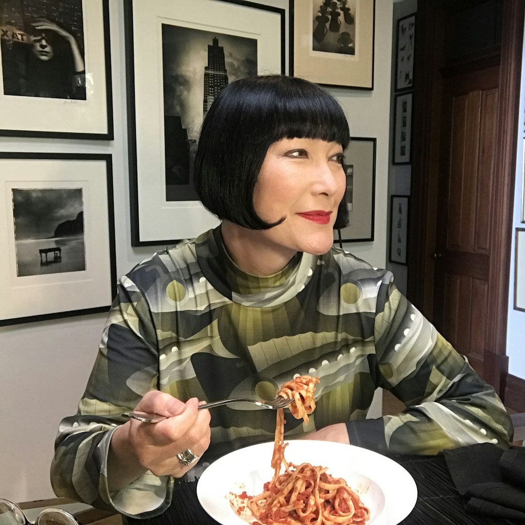 A woman with dark bobbed hair seated at a table lifts a forkful of pasta from a bowl.