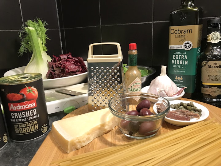 A can of crushed tomatoes, wedge of parmesan cheese, grater, small bowl of black olives, small dish of anchovies and capers, head of garlic, fennel, radicchio and bottles of Tabasco sauce, olive oil and vinegar.
