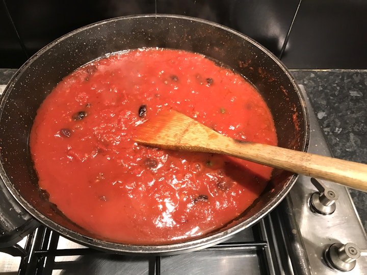 Tomato sauce studded with olives and capers in a frypan, on which a wooden spoon rests.