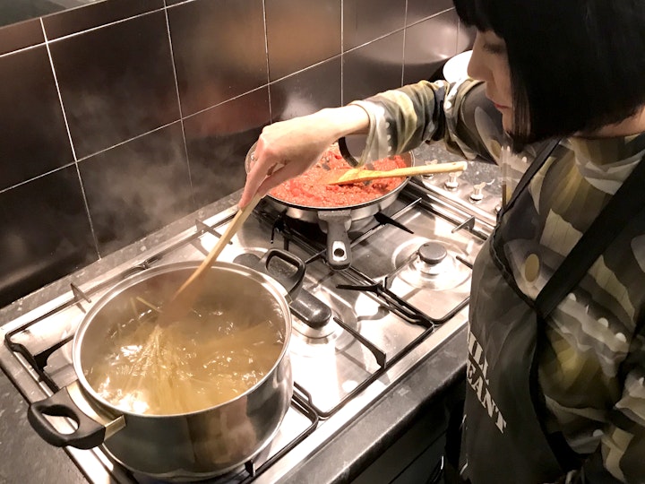 A woman with dark bobbed hair, wearing an apron, stirs pasta in a saucepan on a stove. A tomato-based sauce is in a frypan in the background.