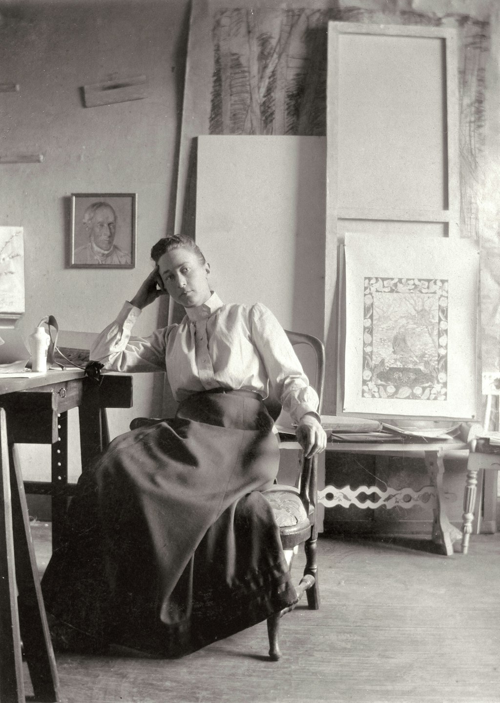 Black-and-white photograph of a woman with hair pulled back in a bun, wearing a long skit and high-necked long-sleeved blouse. She sits facing us, her elbow on a table, with her hand propping up her head. In the room behind her are various artworks, some facing the wall.