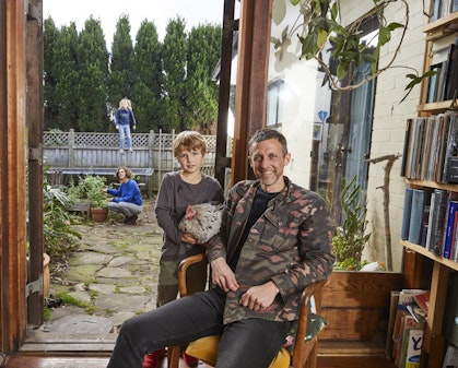 A man sits in a chair, with a young boy standing at his right. Through the door and window behind them we see an adult crouching in a garden and a child jumping on a trampoline.