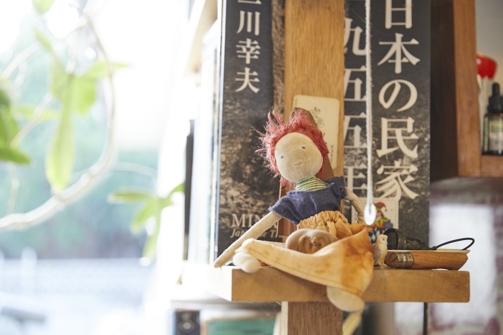 A small handmade doll rests on the end of a wooden shelf, with a publication that has Japanese script on the cover. 