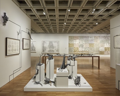 Large white-walled gallery space with drawings hanging on walls and multiple objects on plinths.