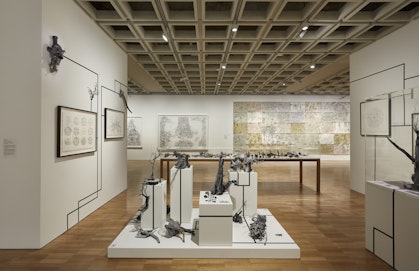 Large white-walled gallery space with drawings hanging on walls and multiple objects on plinths.