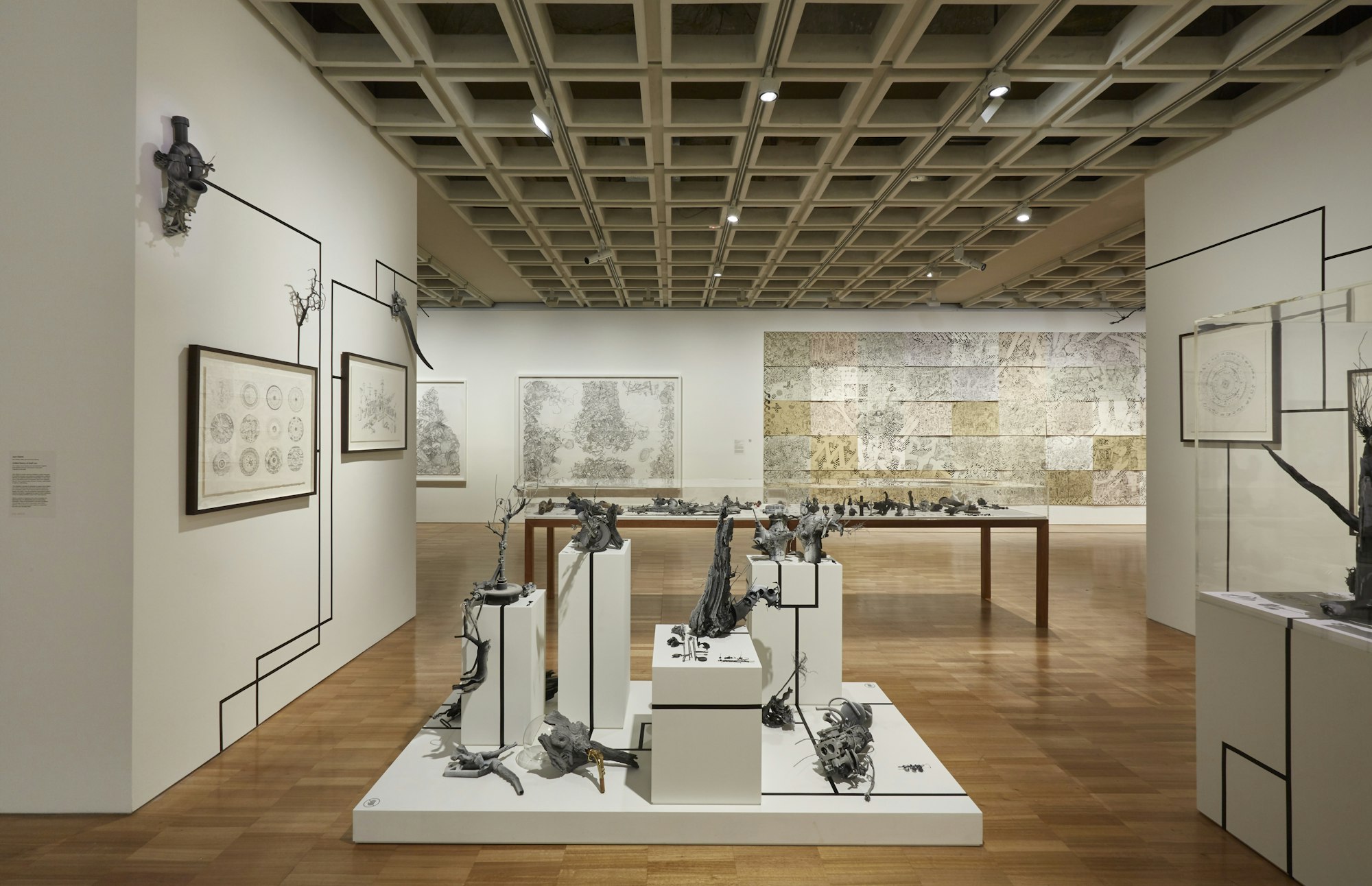 Large white-walled gallery space with drawings hanging on walls and multiple objects on plinths.