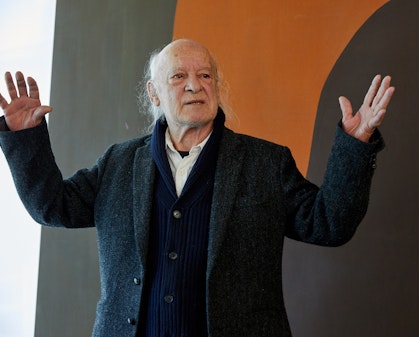 A balding man dressed in a white shirt and dark cardigan and coat in front of an abstract painting with his hands held up as he speaks.