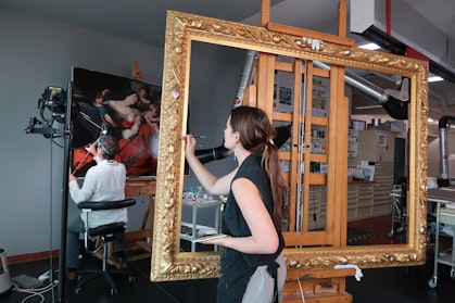 A person works on a painting on an easel while another person works on an ornate gold frame on an easel.