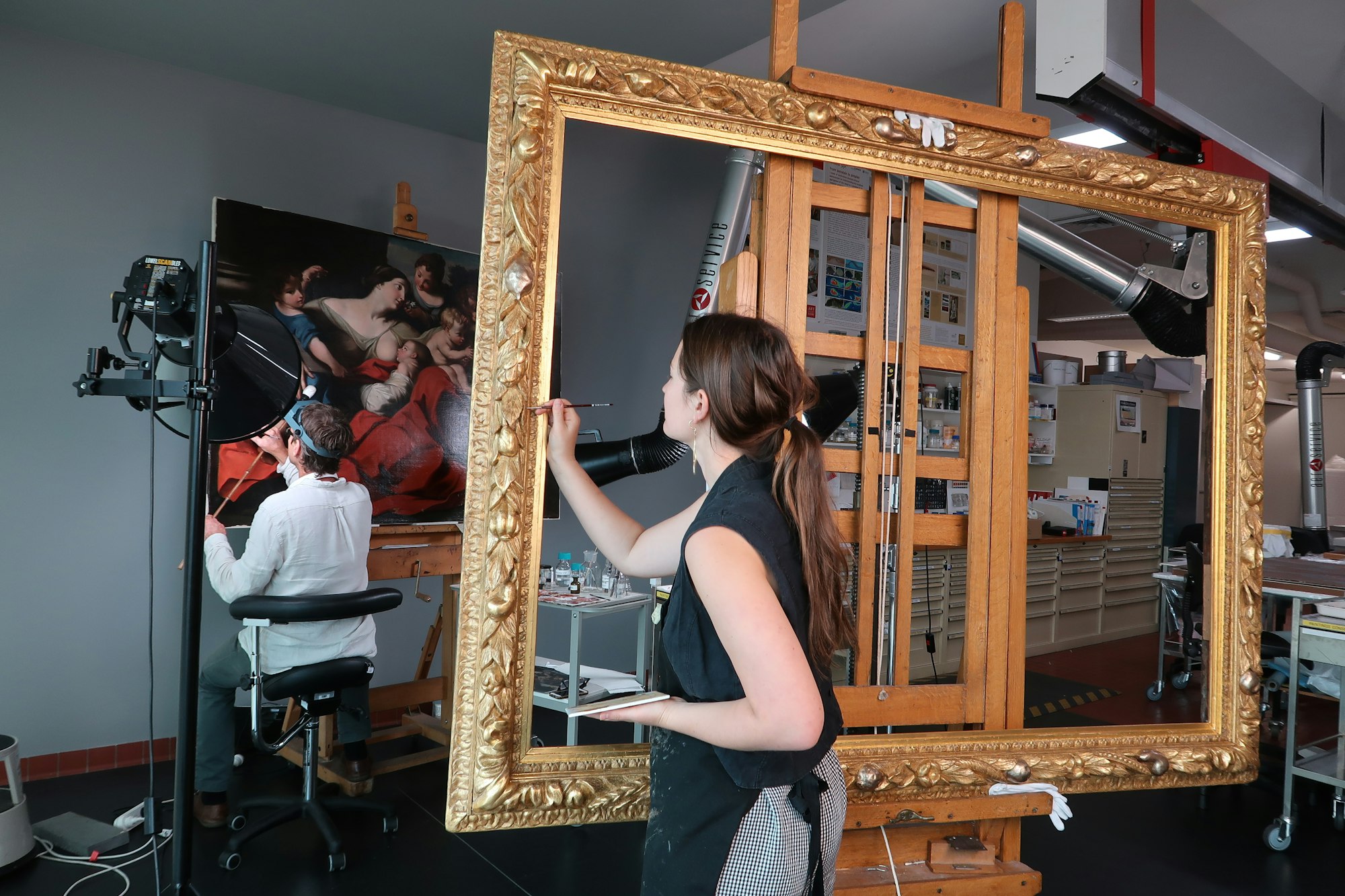 A person works on a painting on an easel while another person works on an ornate gold frame on an easel.