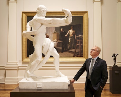 A man wearing a suit, tie and glasses stands looking up at a white sculpture of a man wrestling a huge snake.