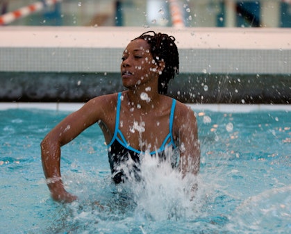 A dark-haired woman dressed in a swimsuit beats the water in a swimming pool with her hands, creating splashes.