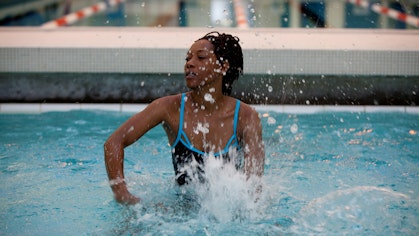 A dark-haired woman dressed in a swimsuit beats the water in a swimming pool with her hands, creating splashes.