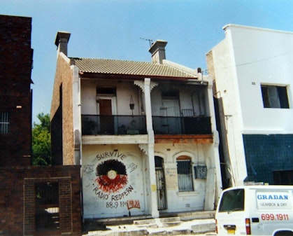 Terrace house in a row of buildings, with a stylised Aboriginal flag painted on the front and the words 'Survive' above and 'Radio Redfern 88.9 FM' below.
