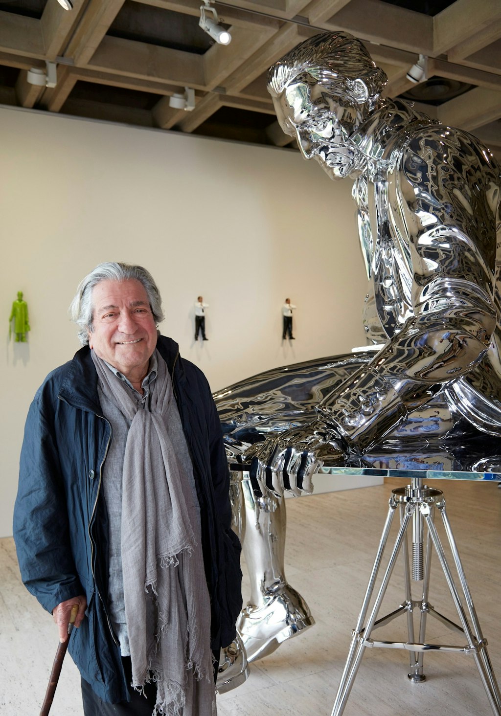 A grey-haired man dressed in a grey scarf and blue jacket leans on a walking stick next to a huge shiny silver statue of a seated figure of explorer James Cook.