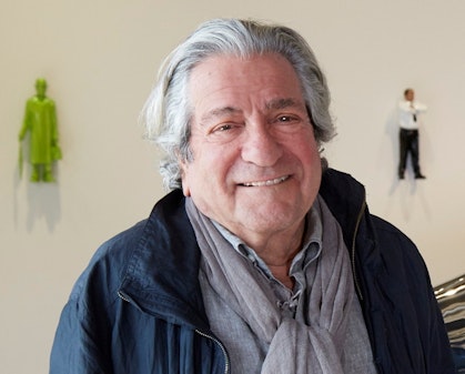 Grey-haired man dressed in grey shit, grey scarf and navy jacket. Two small human figures hang on the wall in the background.