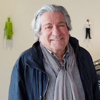 Grey-haired man dressed in grey shit, grey scarf and navy jacket. Two small human figures hang on the wall in the background.