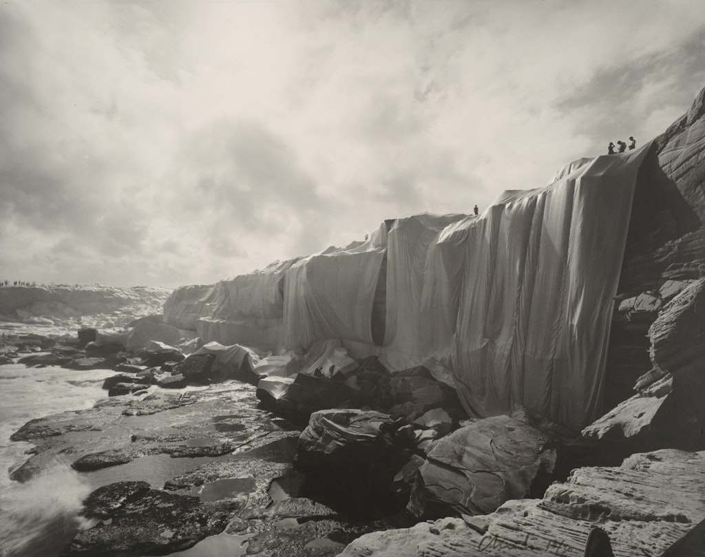 Black-and-white photograph of a coastline with cliffs draped in fabric, on top of which stand a few people.